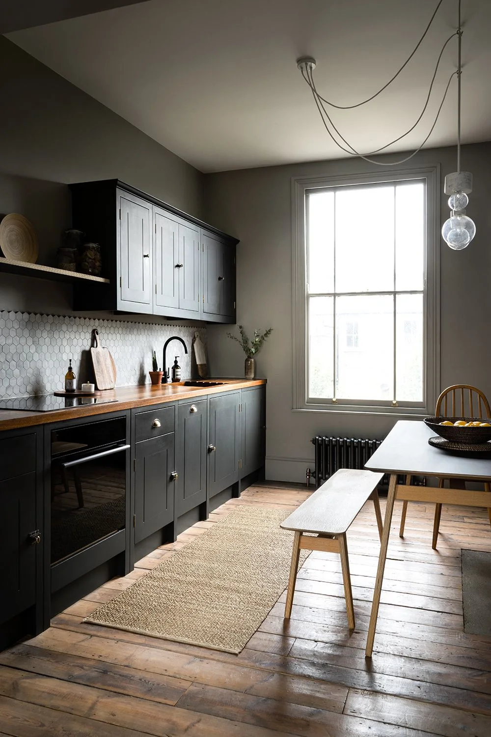 A small kitchen has dark upper and lower cupboards, a warmer wood counter and pale backsplash beside a window, narrow dining table and bench