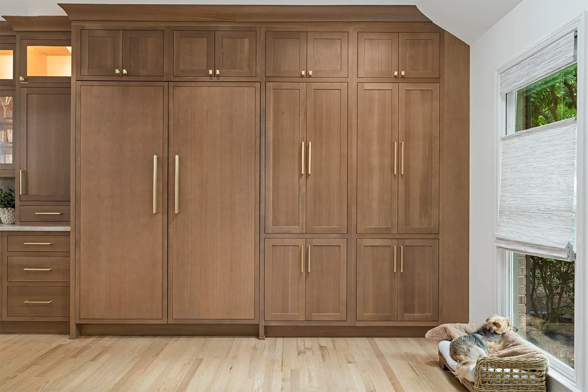 Stained white oak panels conceal large kitchen appliances beneath upper cabinets, with long gold-colored pulls and a window beside them.