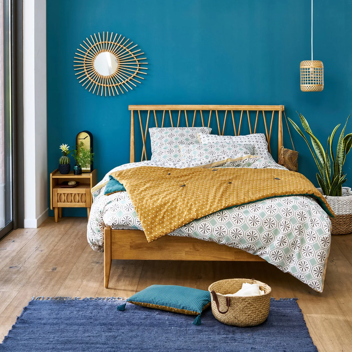 Blue-teal wall behind a wooden bed with patterned white bedding, a mustard quilt and woven accessories.