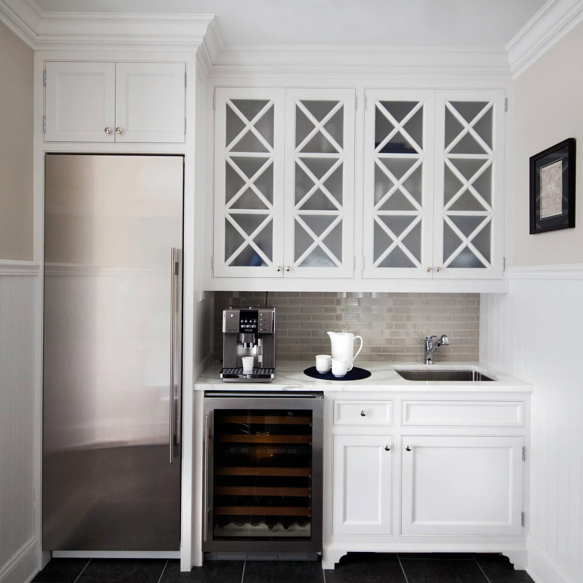 A white two-door cupboard sits above a stainless refrigerator beside glass-front cabinets with crossed muntins.