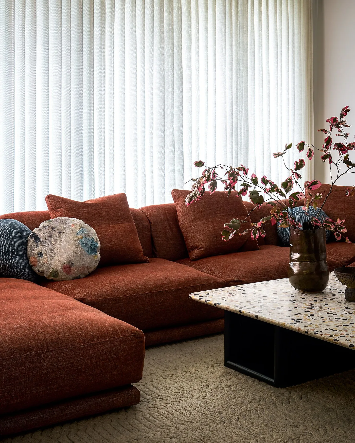 Rust-colored sectional sofa beside long pale curtains and a speckled rectangular coffee table