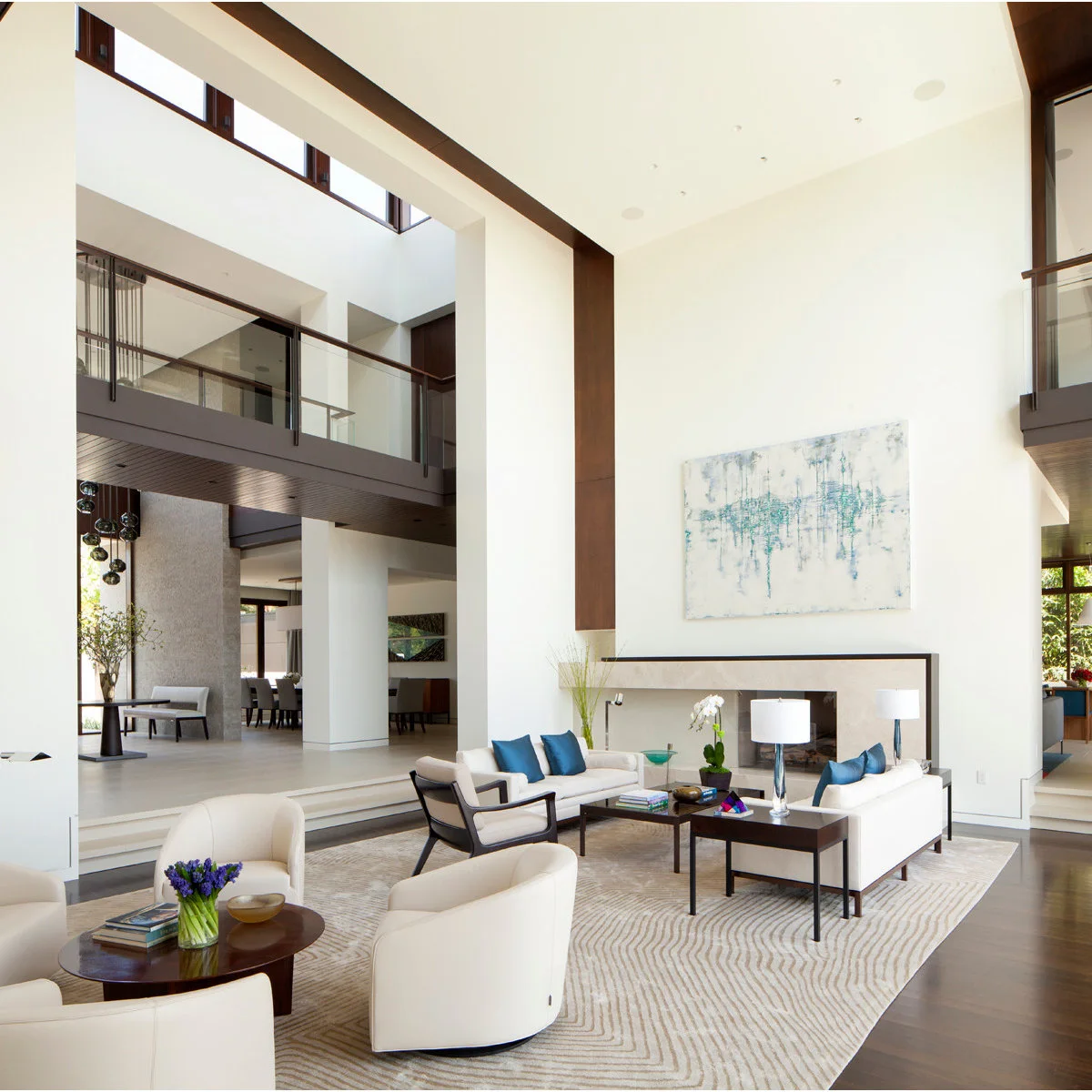 A double-height white room with pale sofas, dark-framed chairs and a large patterned rug below a mezzanine.