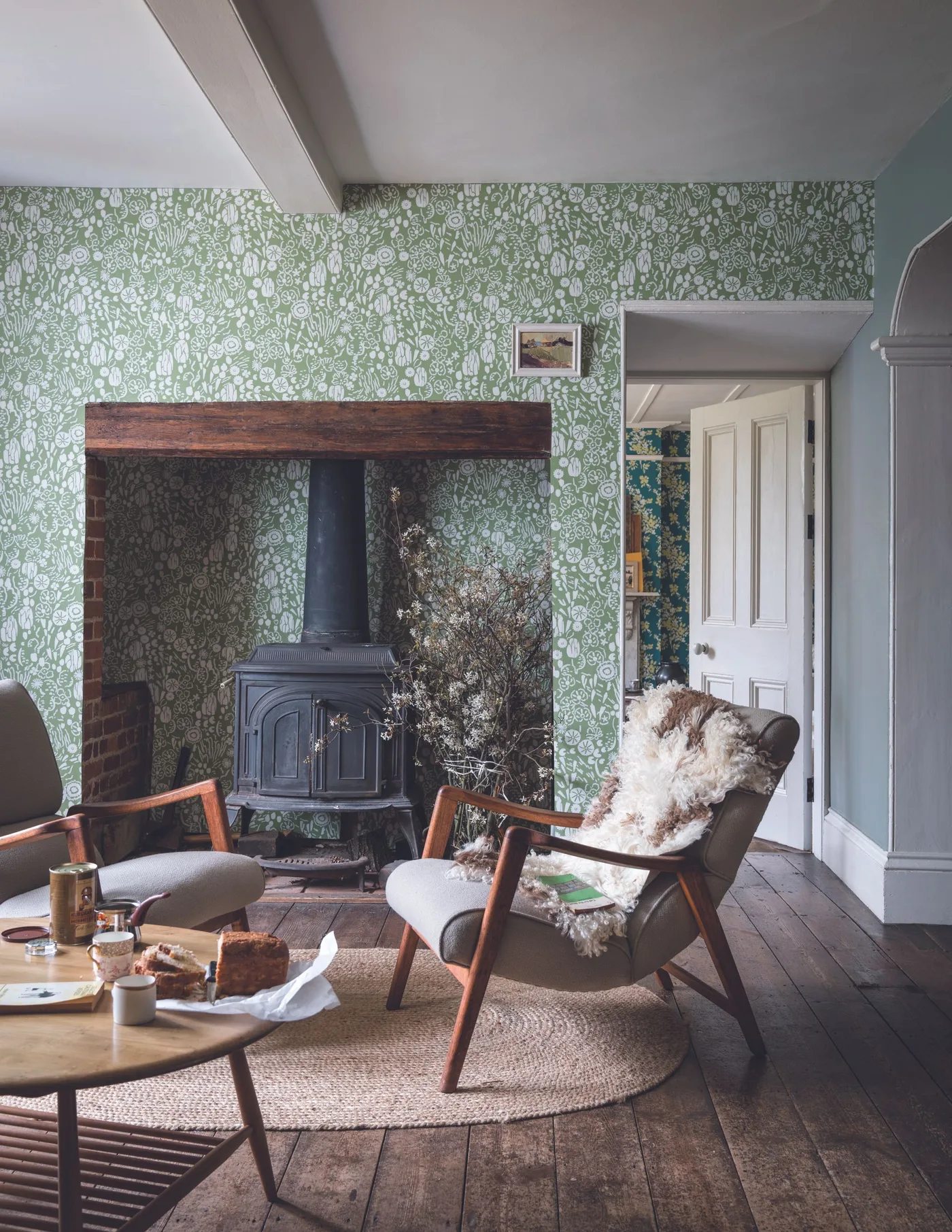 Black stove inside brick opening with broad wooden lintel, green floral wallpaper, simple chairs and round table.