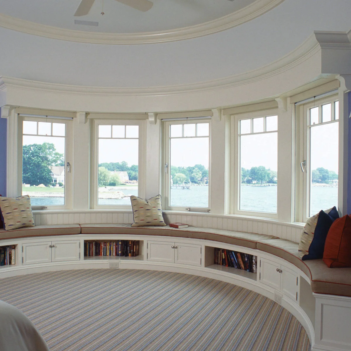 A curved window bench with cream cushions, open book sections and white cupboard doors beneath a water view.