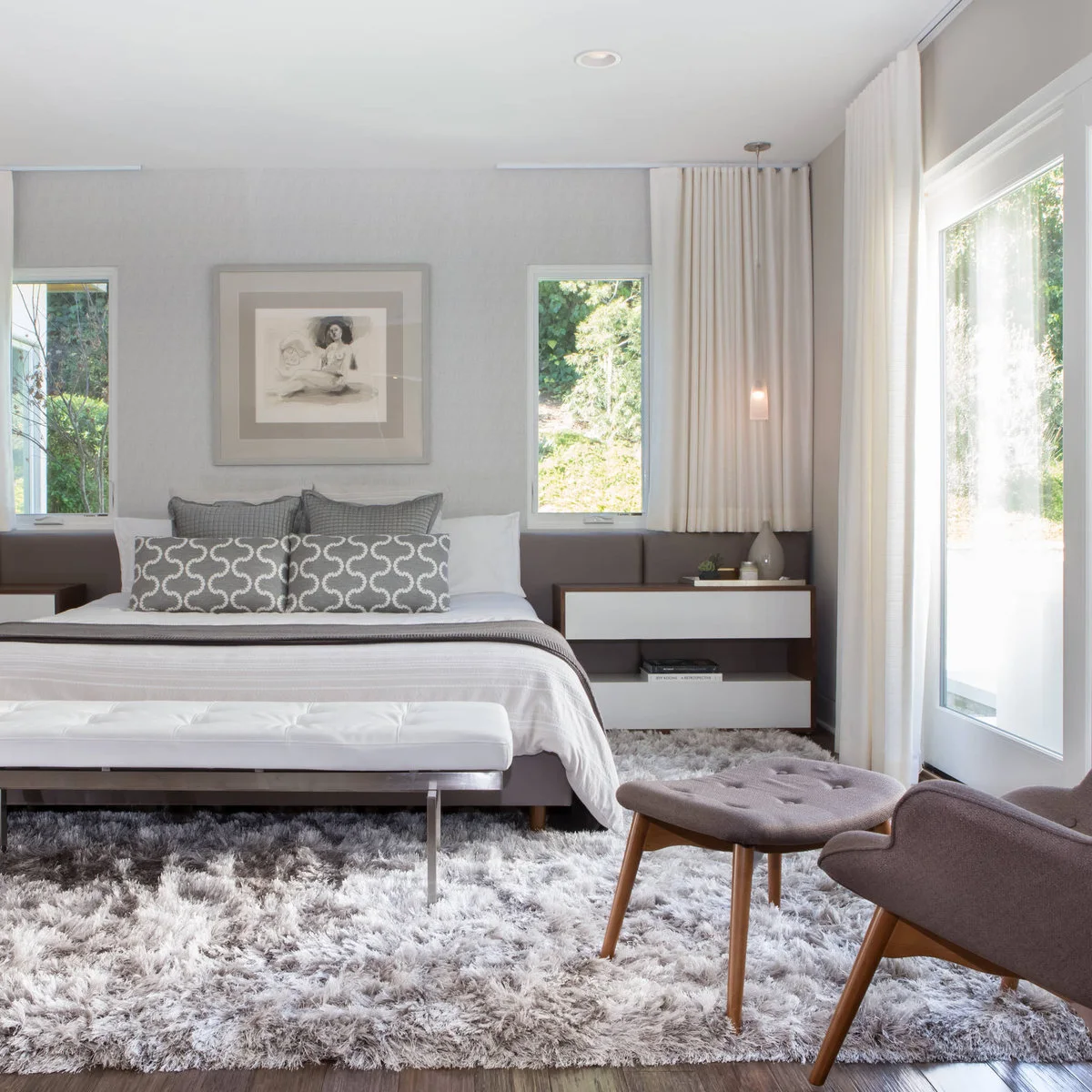 A gray deep-pile rug spans the bed, a white bench and a gray chair with an ottoman beside full-height glazing.