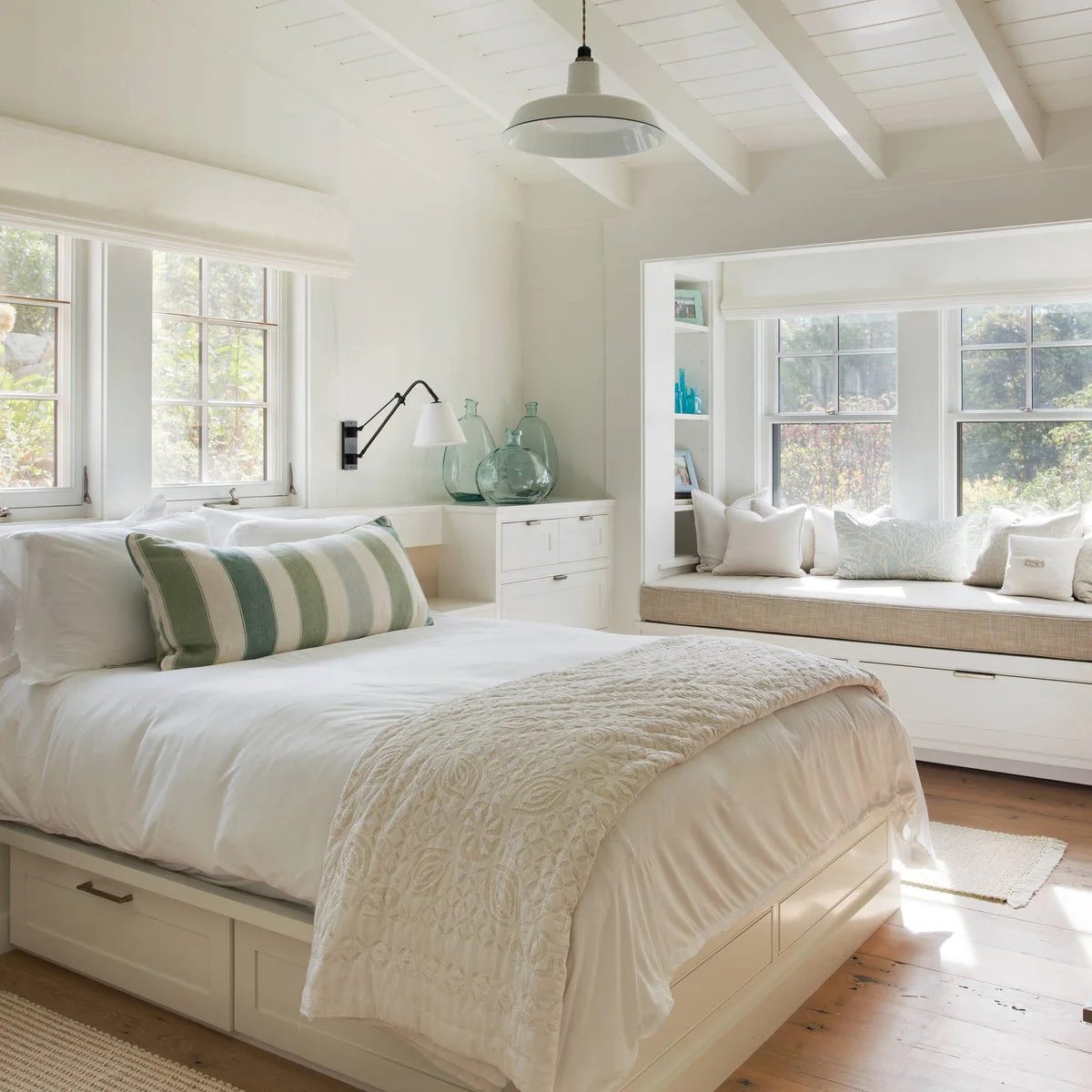 A white bed with drawers below and a matching window bench, beside white fitted storage and an adjustable wall light.