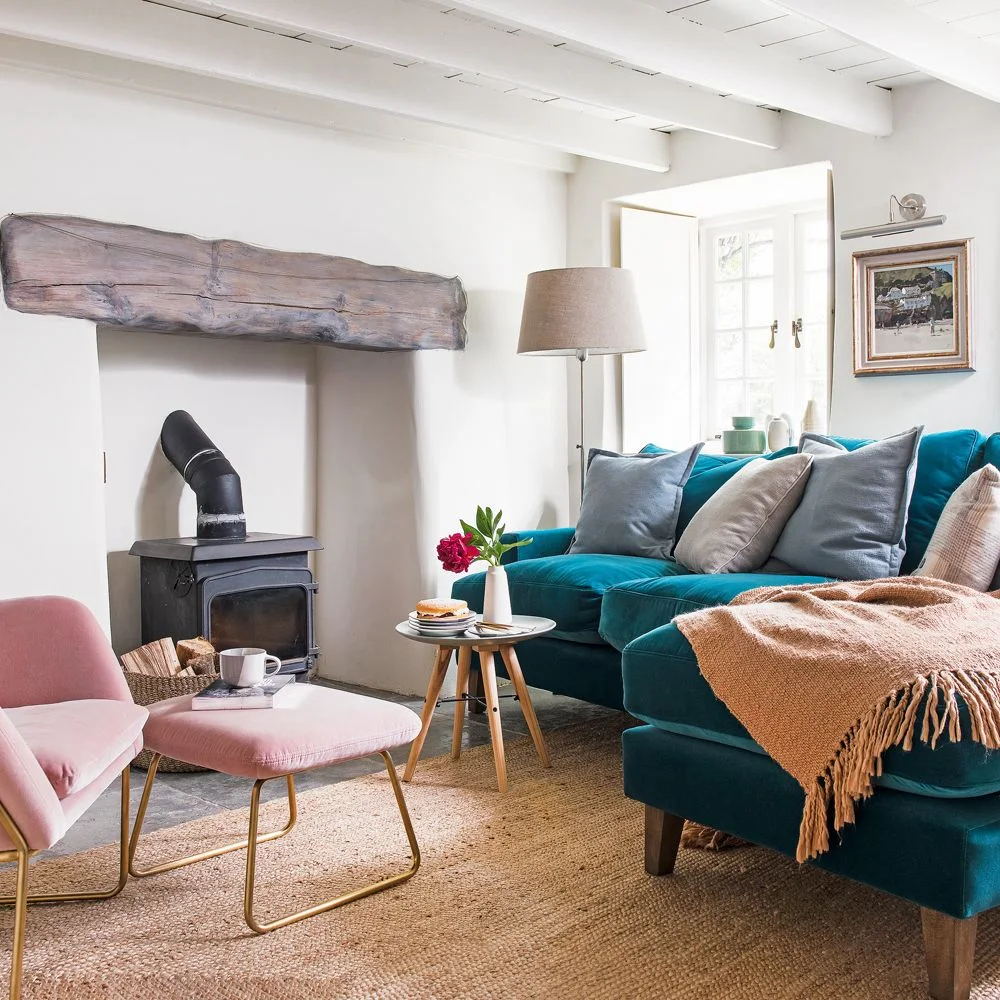 A teal sofa and pink chair sit beside white walls, a broad wood mantel and a small round wood-legged table.