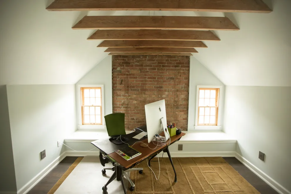A timber desk sits beneath exposed beams between two small windows