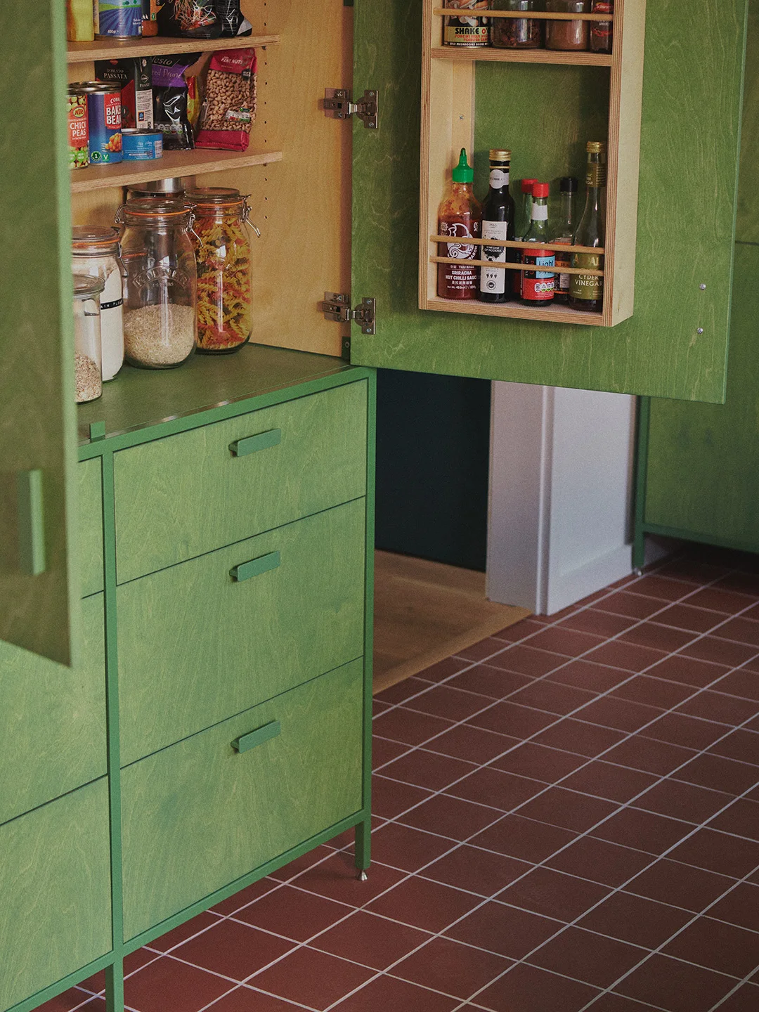 An open green pantry cupboard has wooden interior shelves and a narrow wooden door rack above green drawers