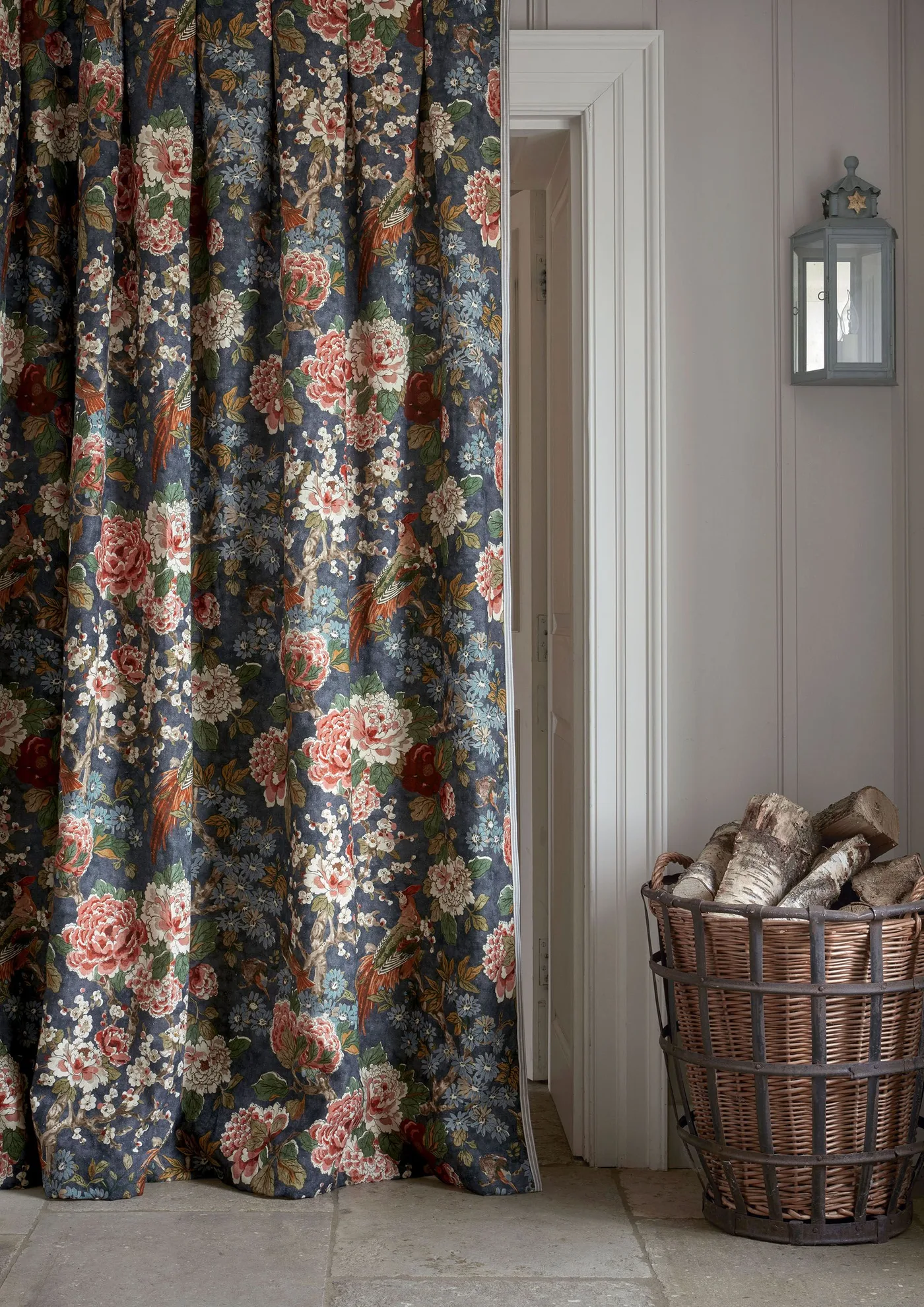 Doorway corner with a dark floral curtain, pale vertical paneling, wall lantern and tall woven basket of logs.