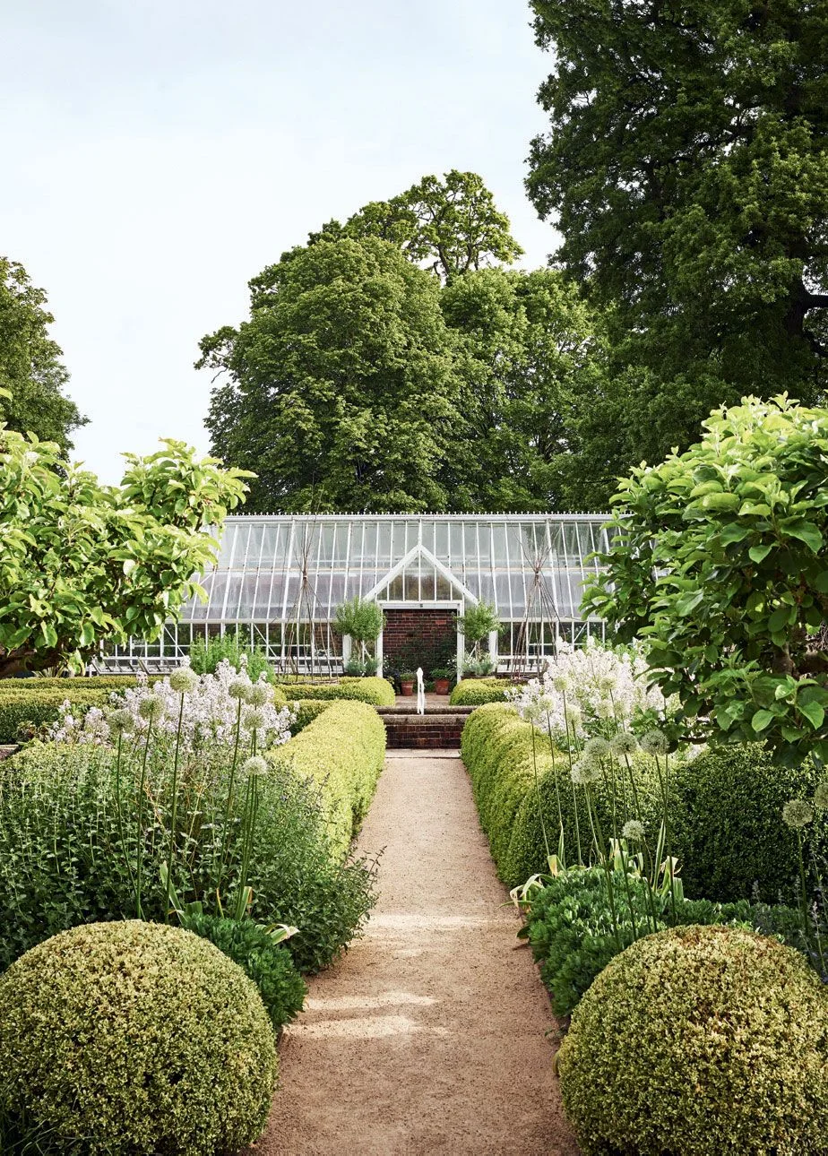 A straight gravel path runs between clipped hedges and flowering plants toward a greenhouse