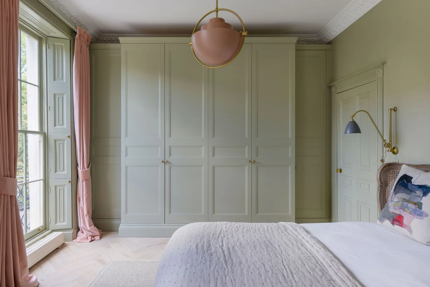 Pale sage paneled wardrobes match the bedroom walls beside pink curtains, pale bedding and a warm-metal pendant.