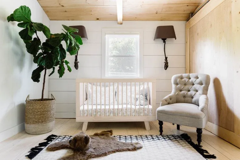 Nursery with pale wooden ceiling and side wall, white crib, upholstered adult chair, woven plant container and patterned rug.