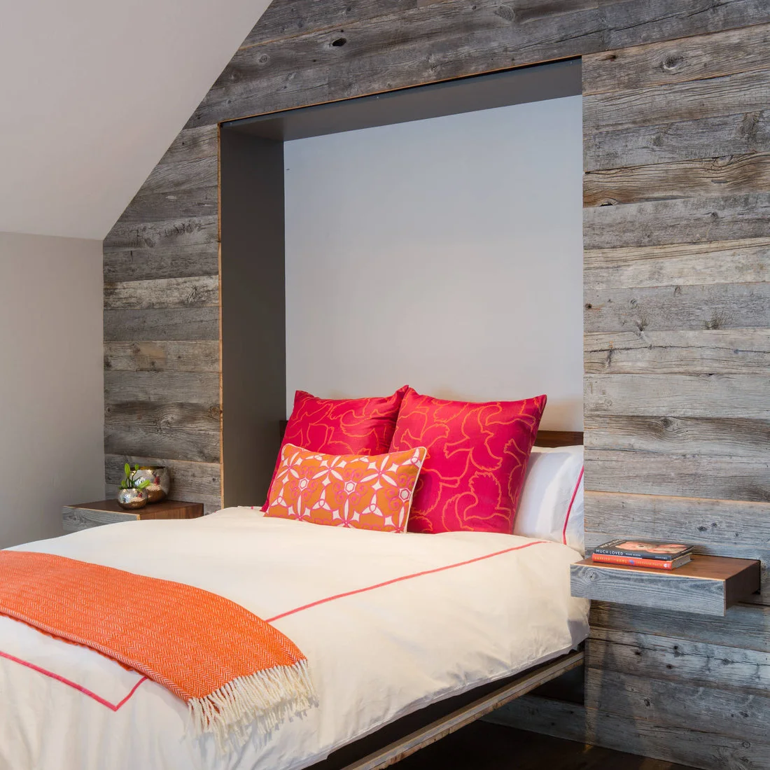 A bed recessed into a gray board wall with floating bedside ledges and orange and pink textiles
