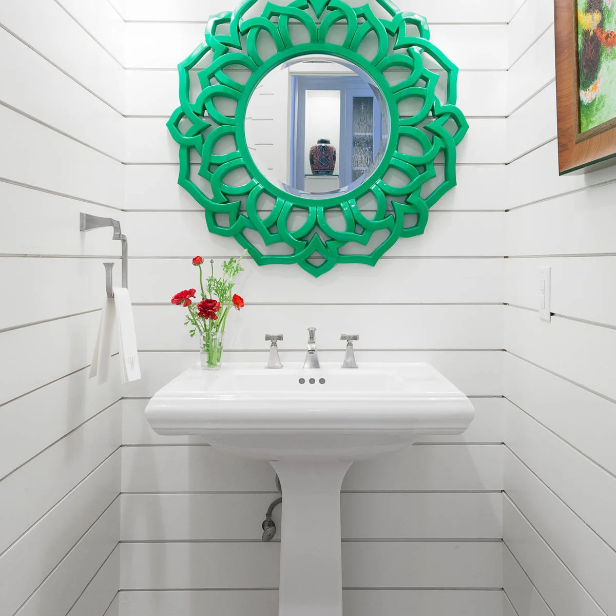 A white pedestal sink stands against horizontal white boards below a bright green round decorative mirror and a small red flower arrangement.