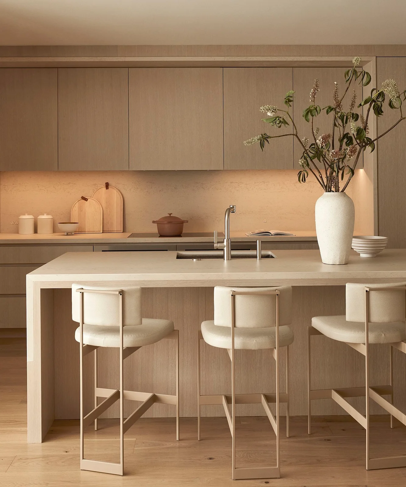 Pale oak flat cabinets and an island sit with cream counters and light upholstered stools beneath warm undercabinet lighting.