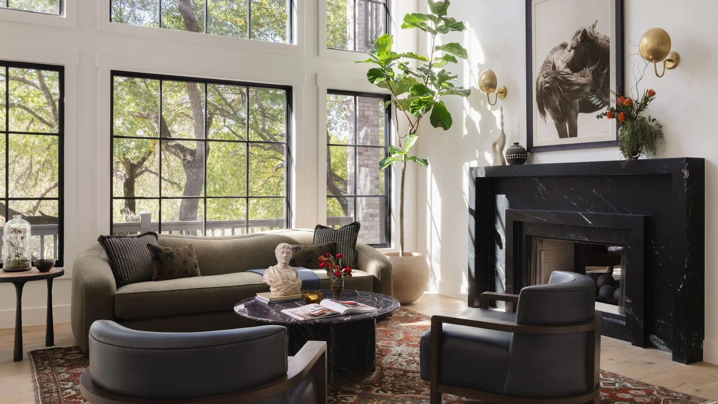 A dark veined fireplace and low table anchor a pale living room with a green-gray sofa, timber-framed chairs and tall windows.