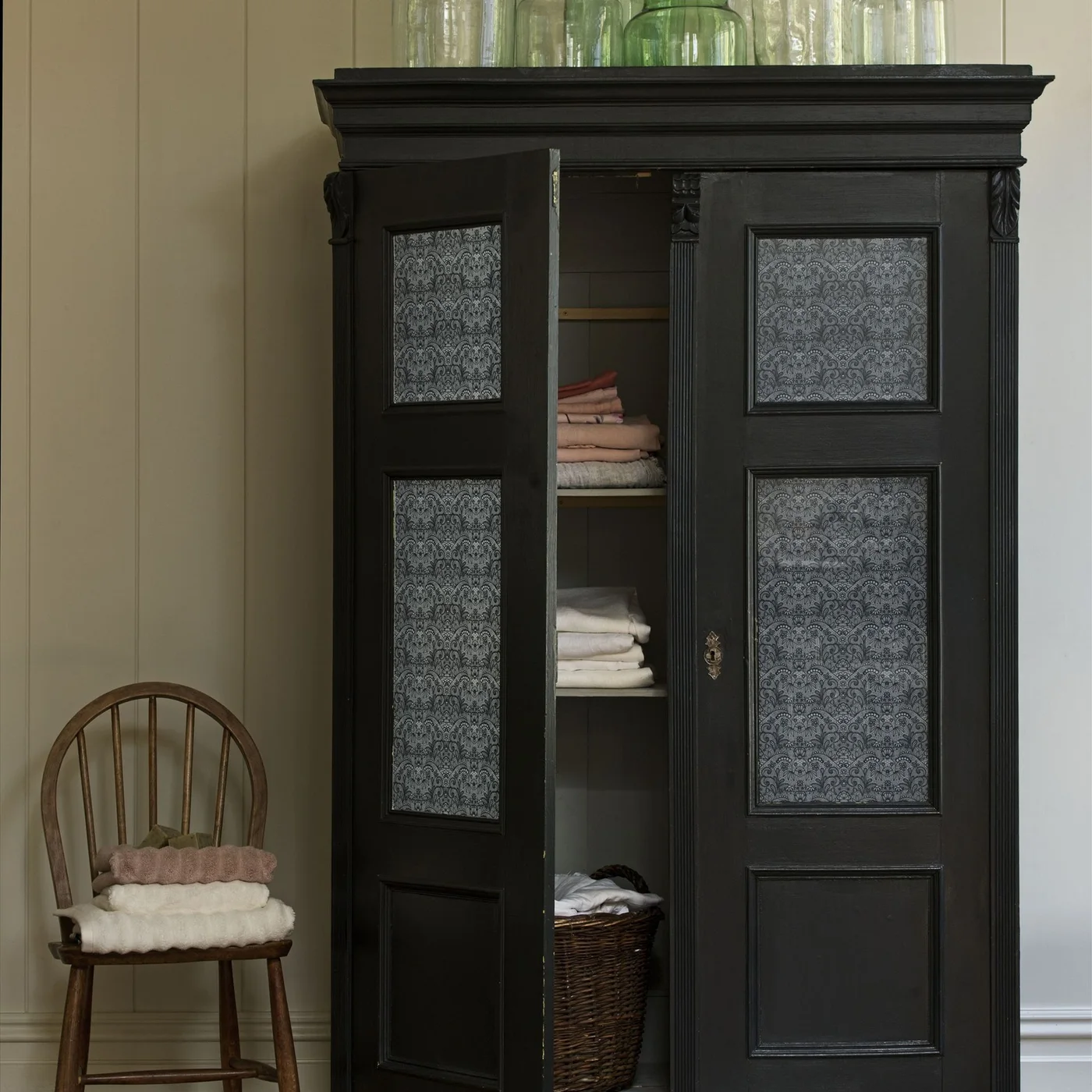 Dark freestanding cupboard with patterned door panels, folded towels, linens and a lower woven basket
