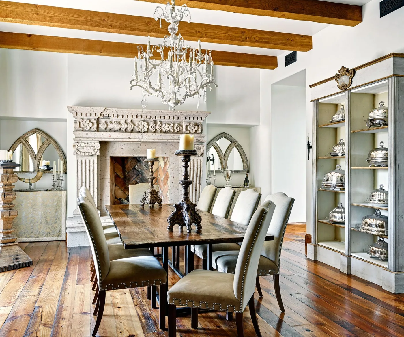 Wooden dining table with pale upholstered chairs, a carved fireplace, exposed beams and glass-fronted storage.