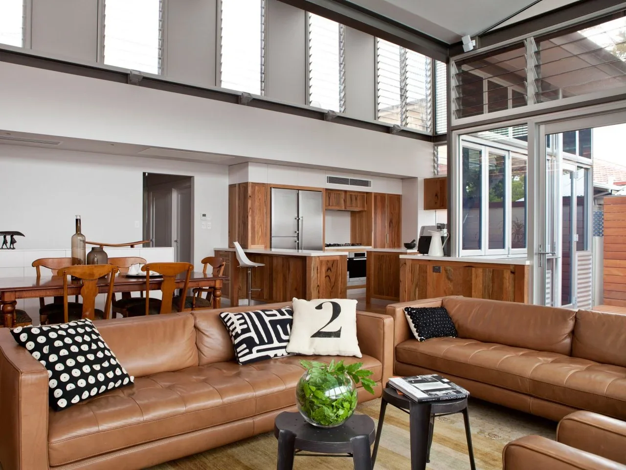 Tan leather sofas sit before timber kitchen cabinets and a timber dining table in a high-ceilinged shared room.