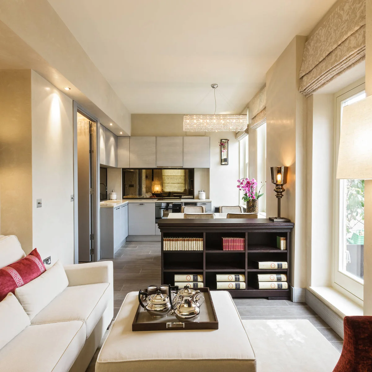 Narrow pale room with a cream sofa and ottoman, low dark bookcase divider, dining chairs and a kitchen at the far end.