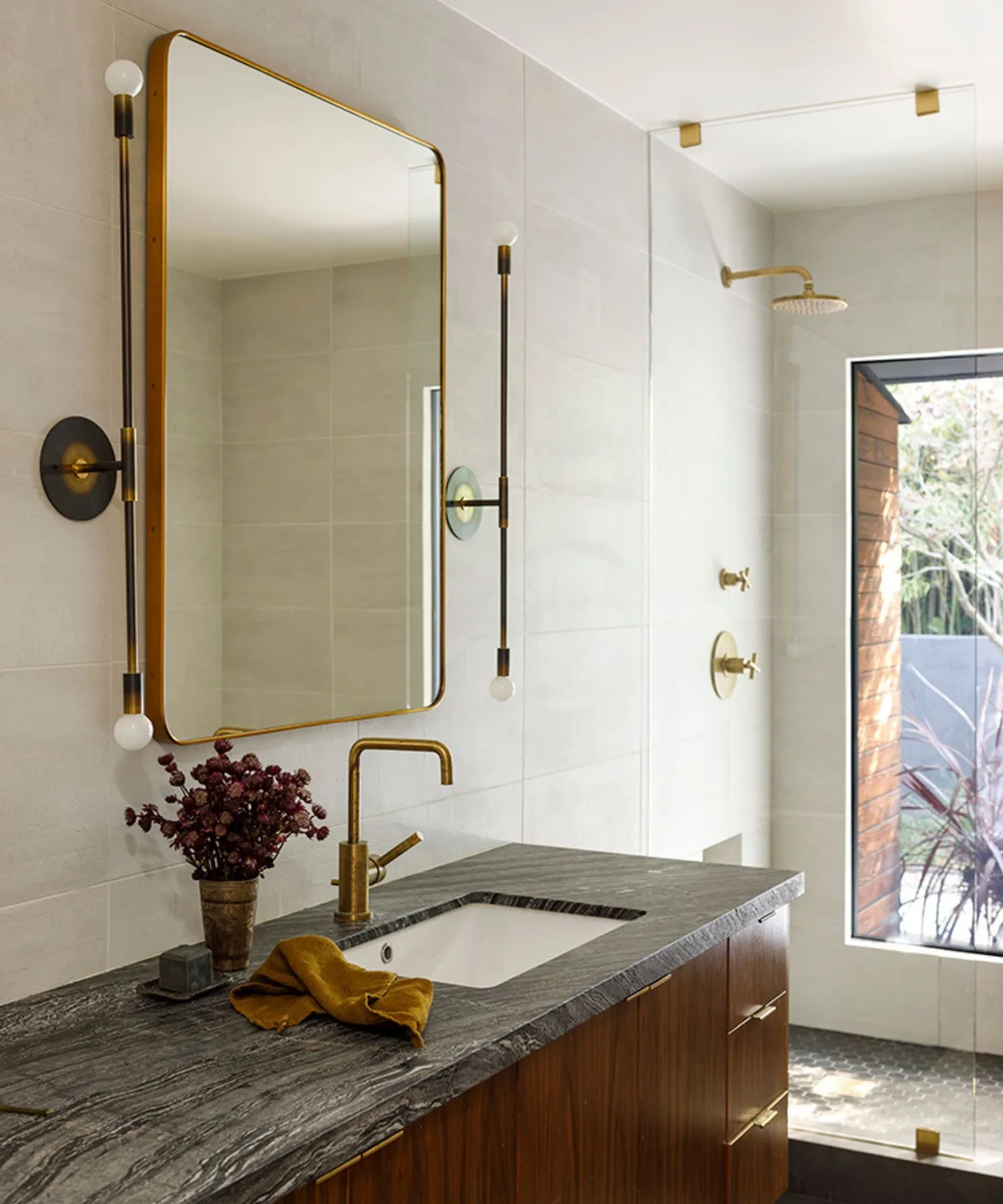 A tall warm metal-framed bathroom mirror sits above a wood vanity and boldly veined counter beside pale tiles, slim globe-ended lights and a shower window