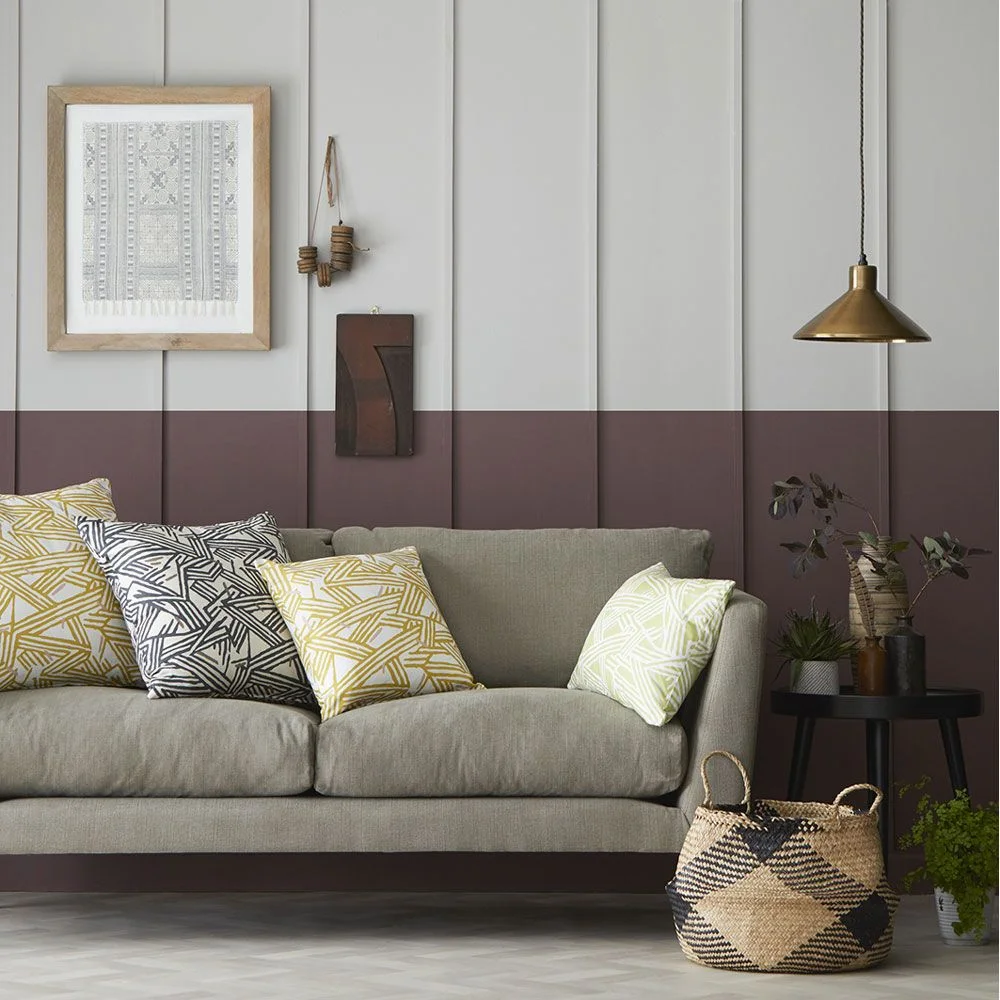 A warm gray sofa with geometric cushions sits beneath pale paneling and a muted plum lower wall beside a woven basket