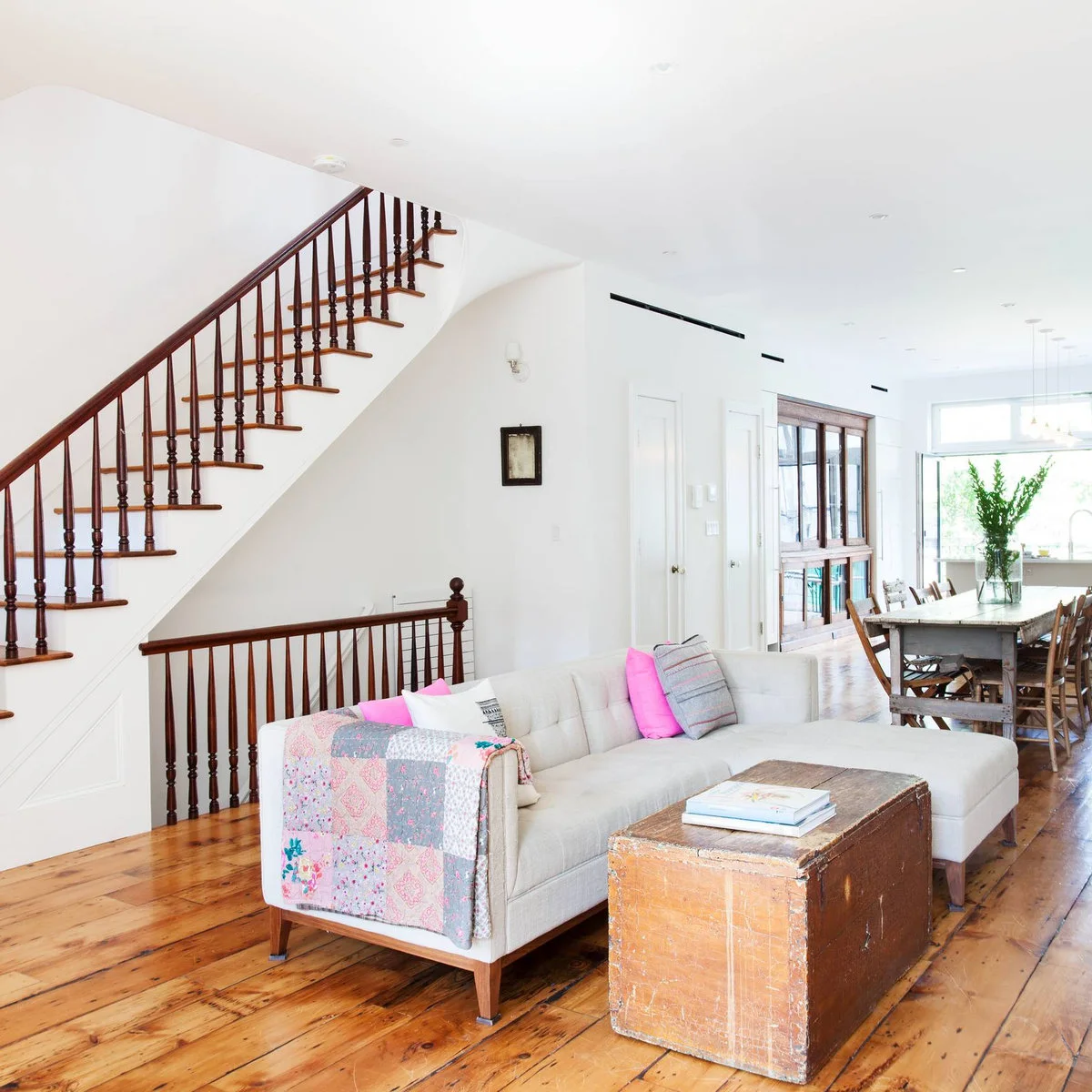Long open room with a pale chaise sofa, wooden trunk coffee table, dining table behind and staircase along one wall.