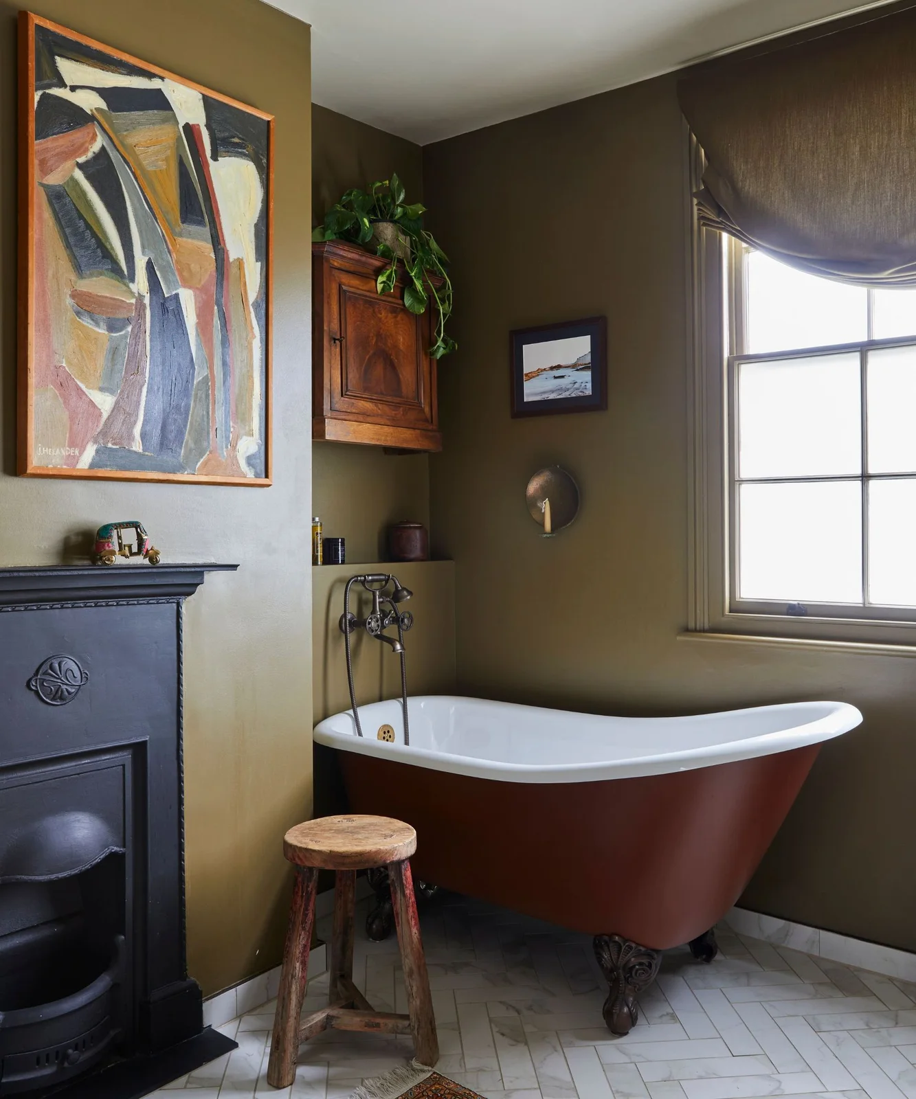 An olive-brown bathroom contains a rust-red clawfoot bath, dark fireplace, framed pictures and pale herringbone flooring.