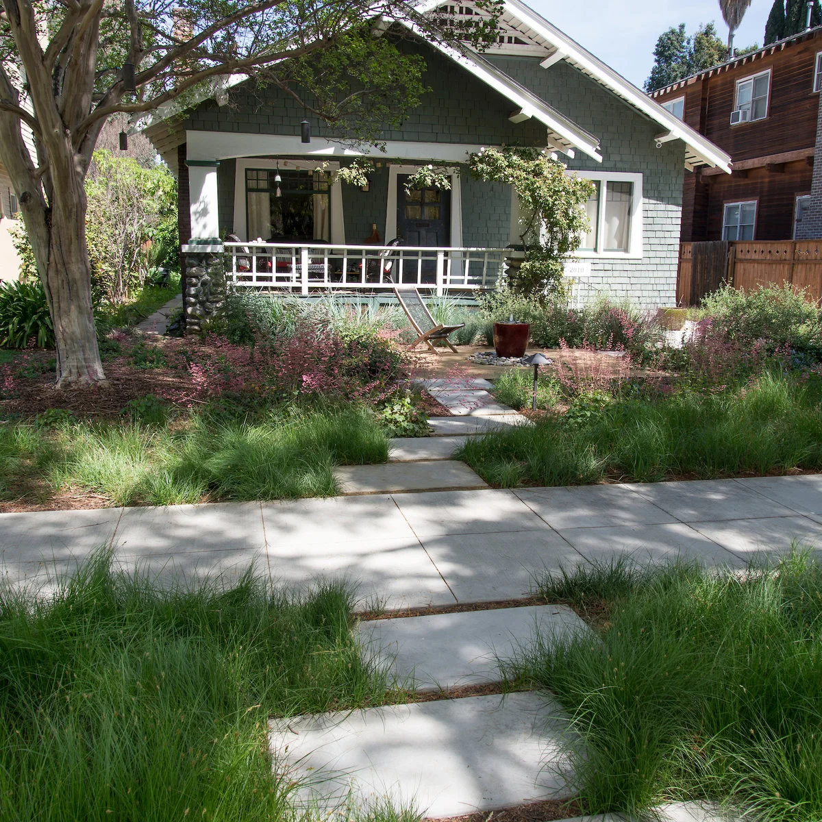 A path crosses meadow-like planting toward a craftsman porch, with a wood chair and large pot beside the approach.
