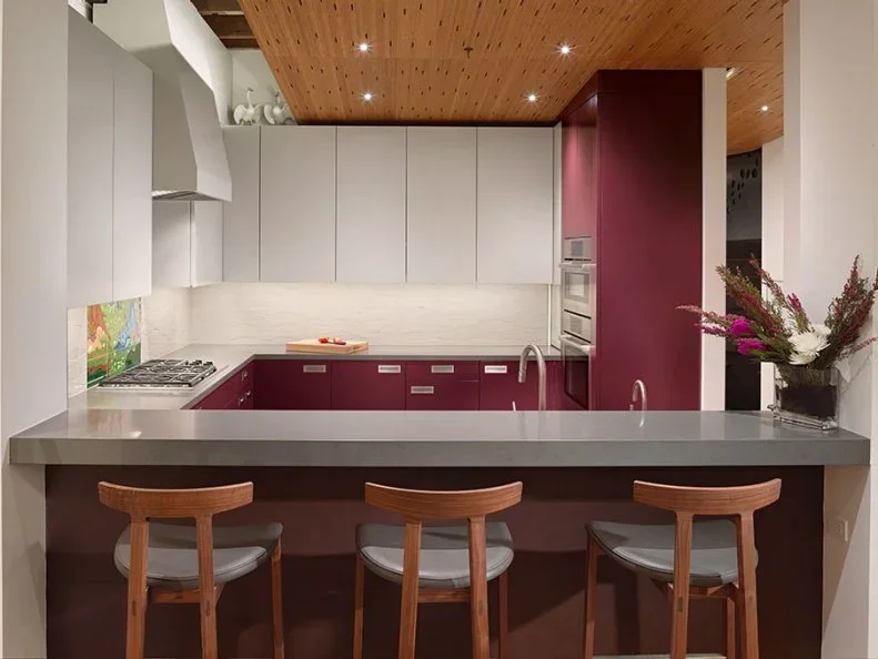 Burgundy lower and tall cabinets sit beneath white wall cupboards in a compact kitchen with a wood ceiling and a gray peninsula counter.