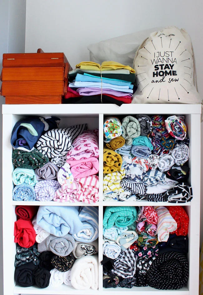 Rolled and folded fabrics fill four white storage cubes, with a wood sewing box and a small fabric stack on top.