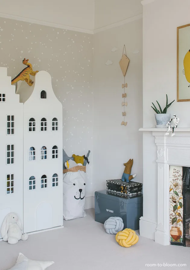 Pale children’s room corner with a house-shaped cupboard, small white sky motifs and toy storage beside a fireplace.