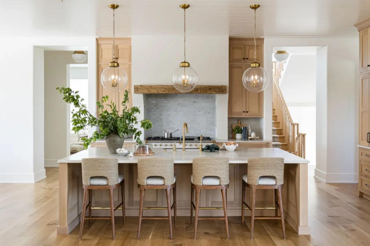Light wood cabinets and a paneled island sit beneath glass pendants, with a pale hood edged by a rustic wood beam.