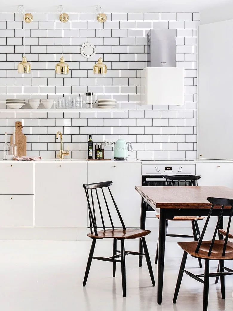 A white kitchen has a broad rectangular tiled wall with dark grout, small warm metal pendant lights, plain fronts and a wood dining table with dark chairs.