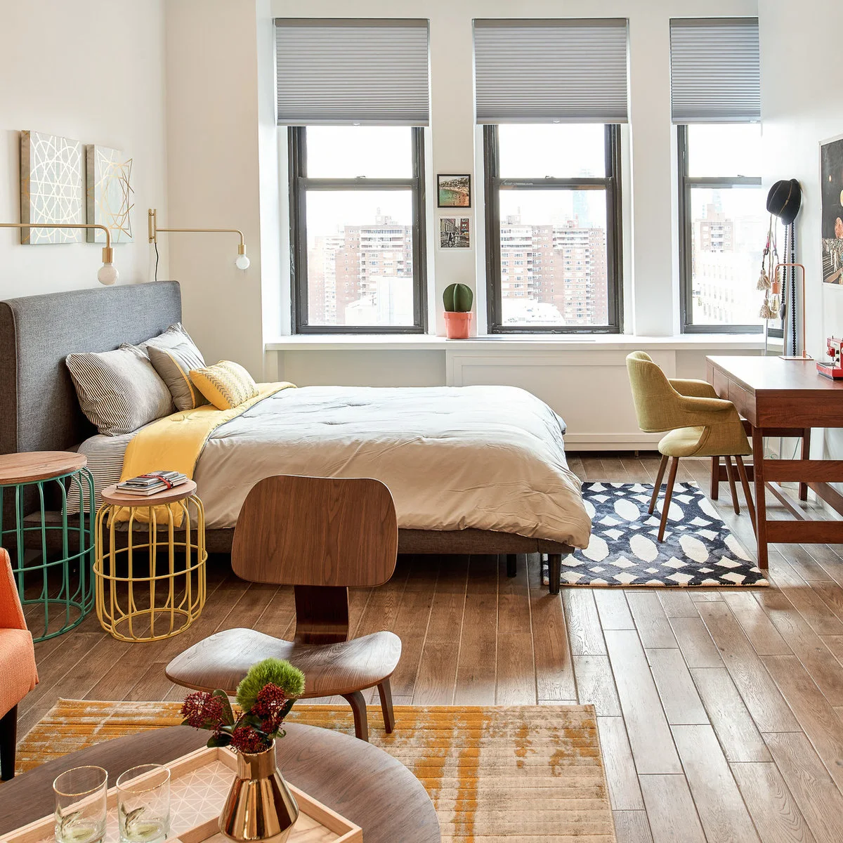 Studio sleeping area with a grey bed, yellow accents, small round tables and a desk by the windows