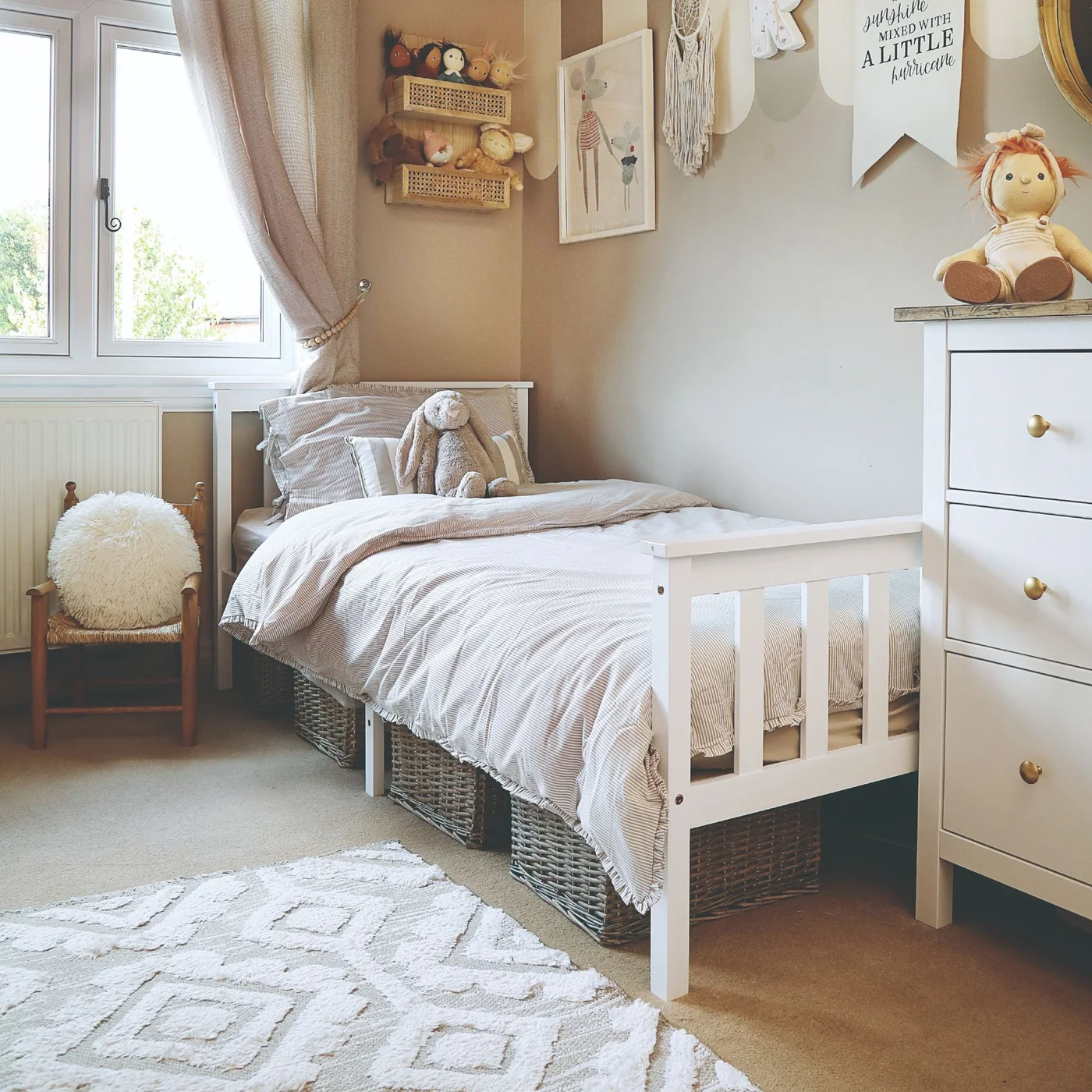 A white single bed has woven baskets beneath it beside a white chest of drawers, pale walls and a light patterned rug