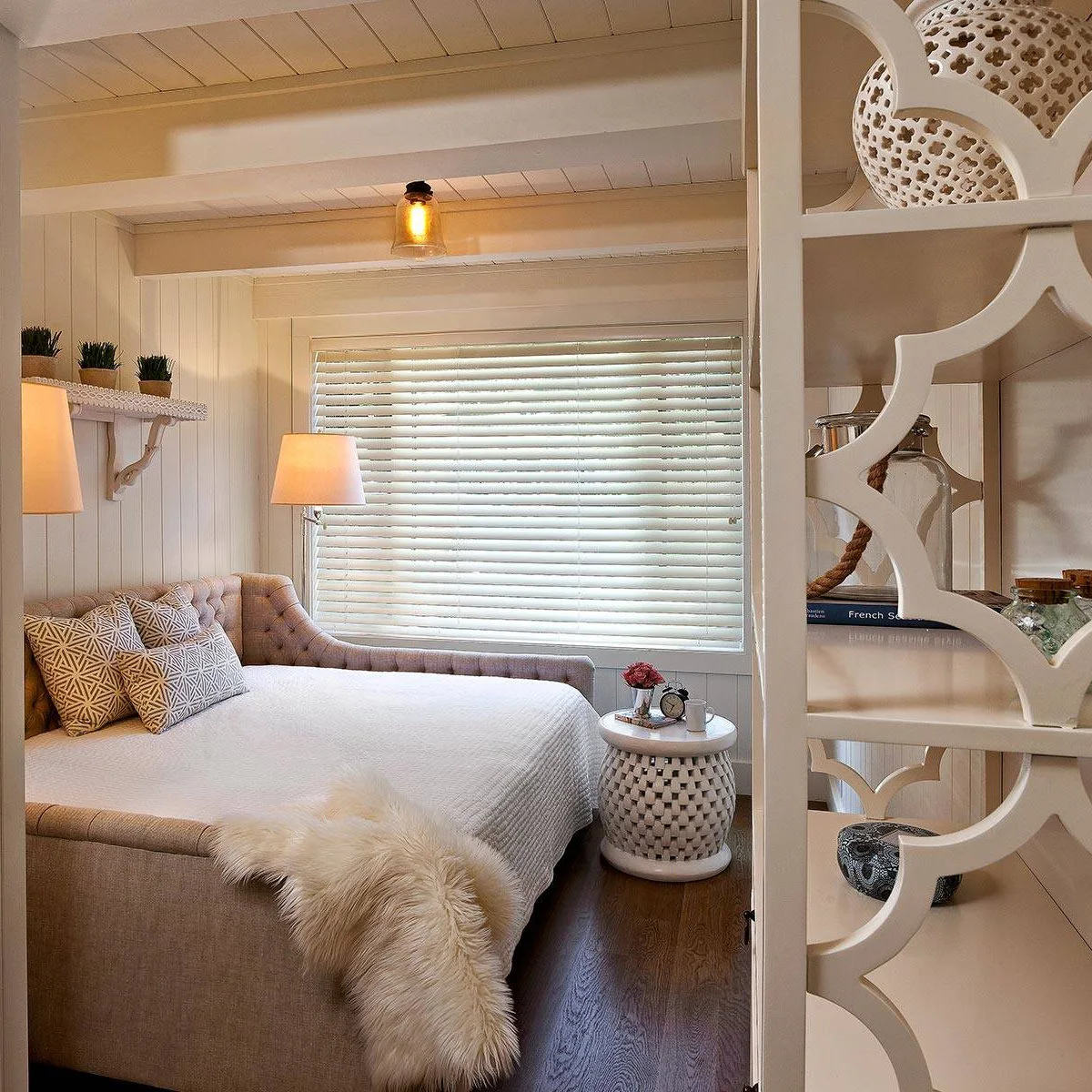 A pale tufted daybed with curved arms fills a small paneled guest nook beside a round white table.