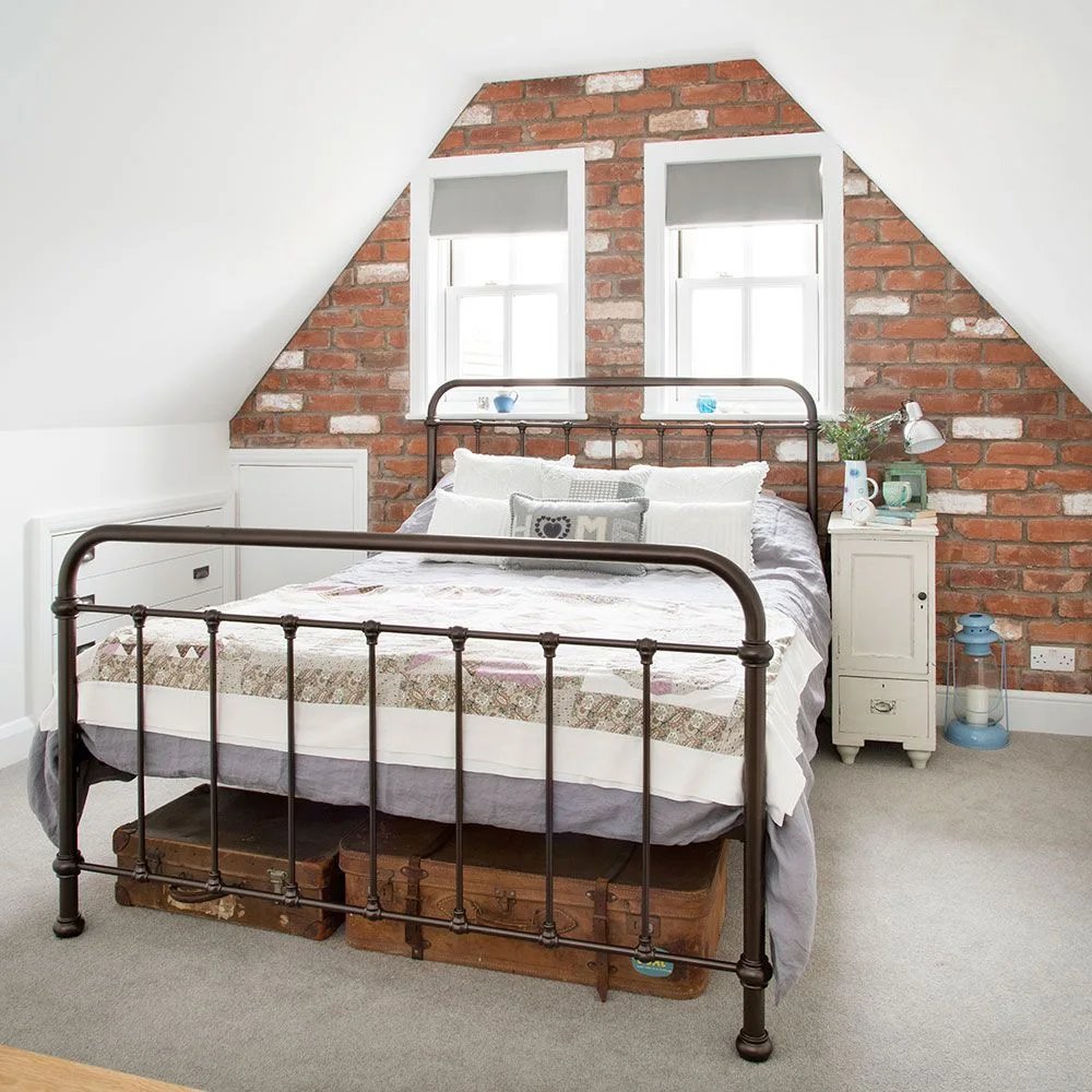 Metal bed below two windows in a brick gable, with low white cupboards and suitcases underneath the bed.