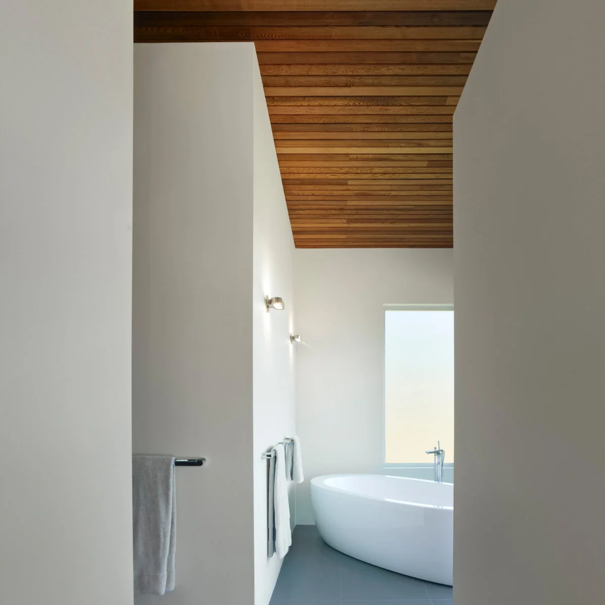 Pale wood plank ceiling over a freestanding bath in a narrow white bathroom