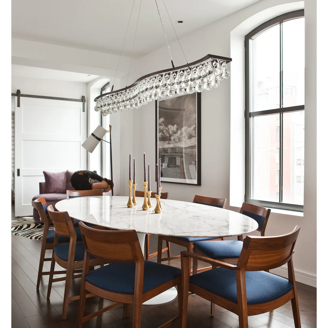 Pale veined oval table with wood chairs and blue seat cushions beneath a long crystal-like pendant