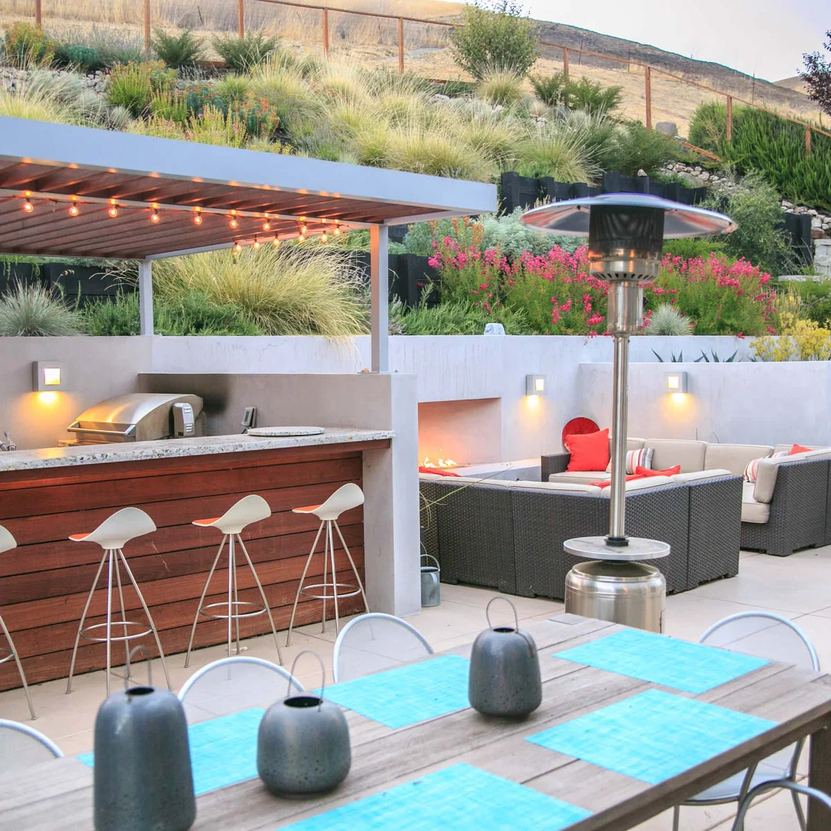 Outdoor bar with horizontal reddish wood cladding, white saddle stools and a planted slope behind the patio