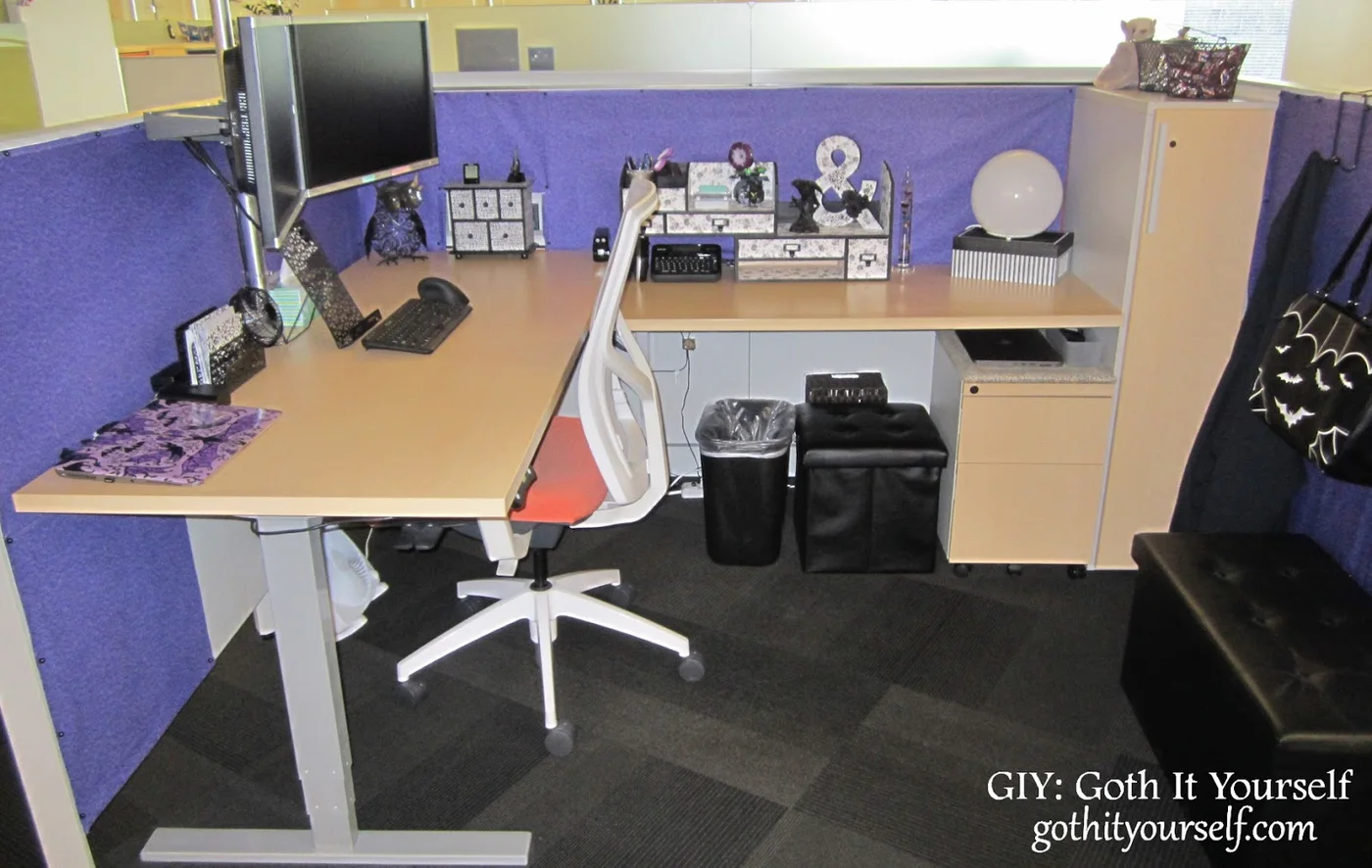 Purple fabric-covered cubicle partitions around an L-shaped desk with black and white organizers, two monitors and a white chair.