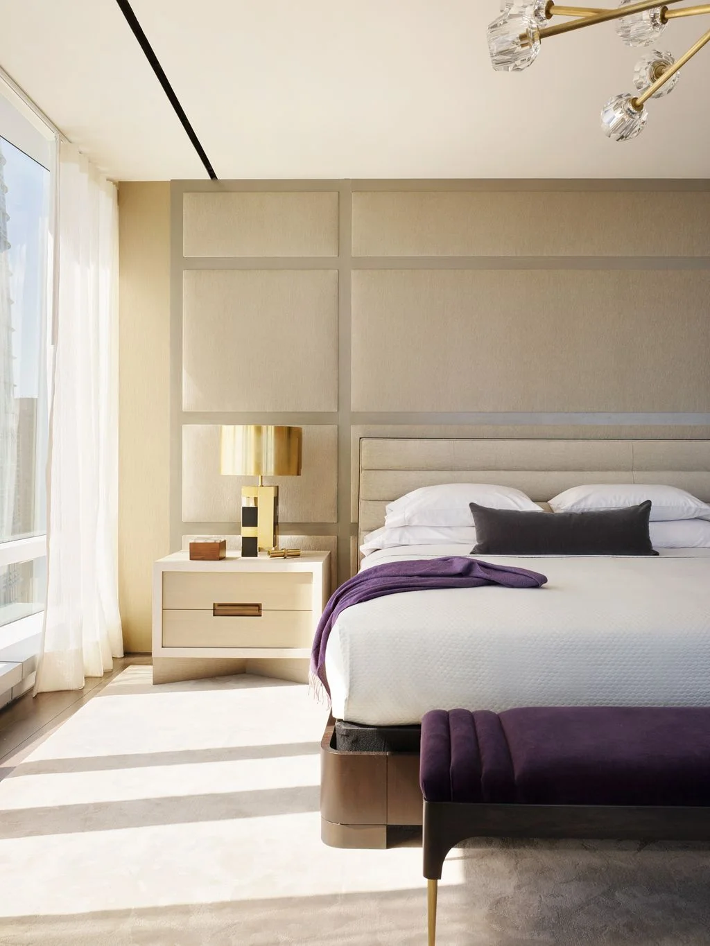 A pale upholstered bed sits against large pale wall panels beside a white drawer unit, a gold-toned lamp and a purple upholstered bench