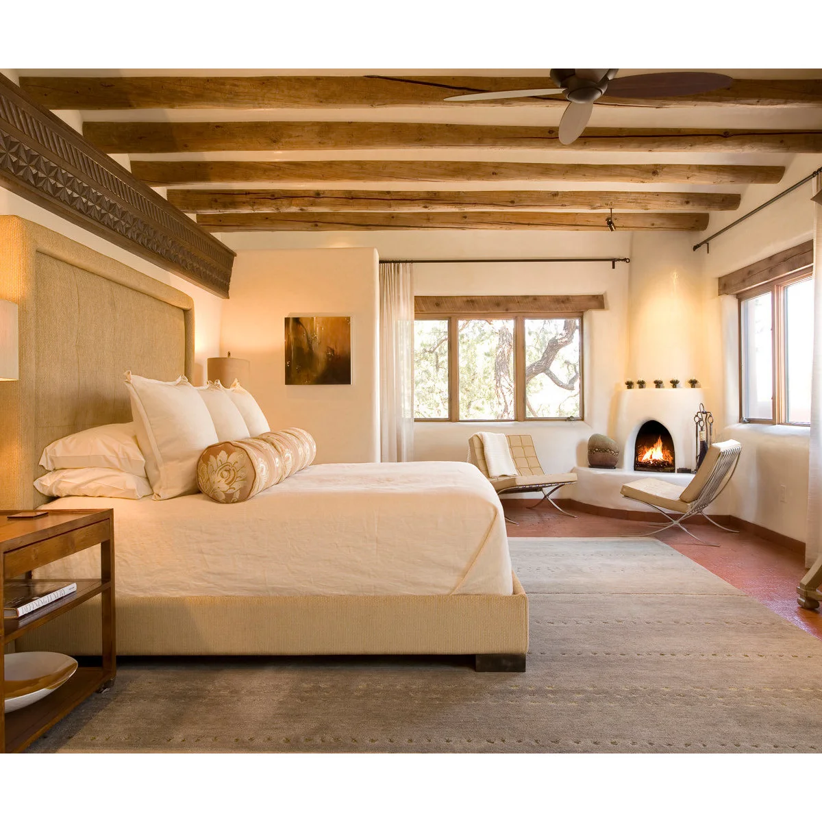 Pale bedroom with rounded corner fireplace, exposed ceiling beams, a large upholstered bed and two light chairs