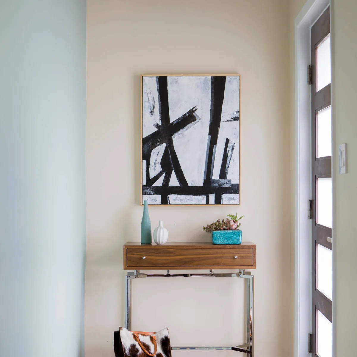 A compact wood-fronted console with fine metal legs beneath a large black-and-white artwork, with small blue-and-white vessels.