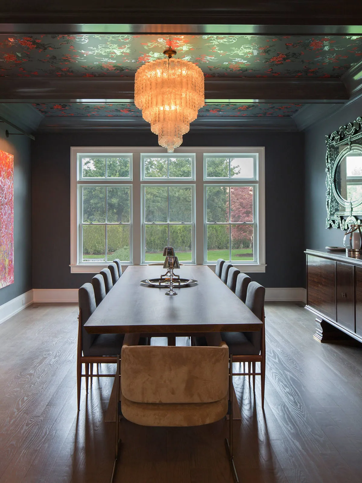 Dark dining room with floral wallpaper in ceiling panels above a long timber table and warm pendant