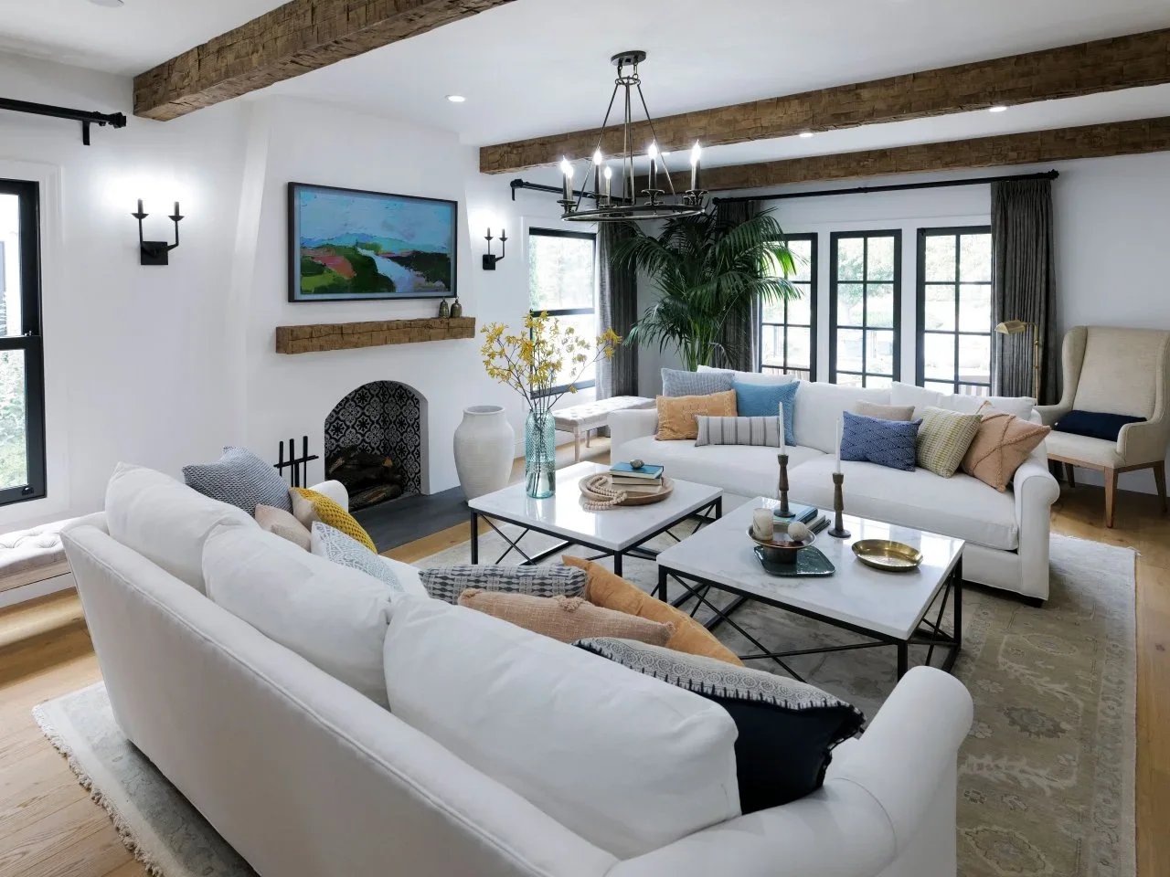 Two white sofas face paired pale-topped coffee tables beneath timber beams and dark window frames.