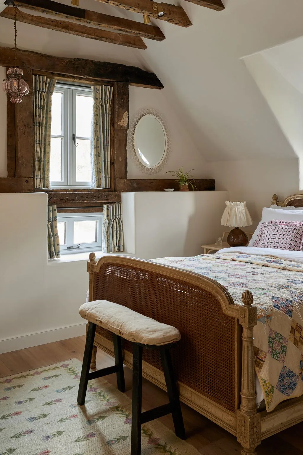 Bedroom with exposed beams, pale walls, woven wood bed frame, patchwork quilt, striped curtains and a small padded stool