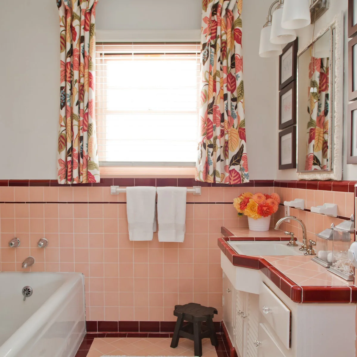 Pink square tile and burgundy trim frame a white bath and tiled vanity beneath colorful floral curtains.